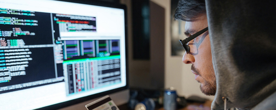A hacker in front of a computer monitor with screens of code visible, inputting credit card info.